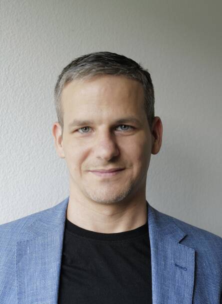 Porträt von Lucas Arquint, einem weissen, 38-jährigen Mann mit kurzen Haaren, vor einer weissen Wand. Er schaut in die Kamera, trägt ein schwarzes T-Shirt und ein Jackett mit feinem Karo-Muster.